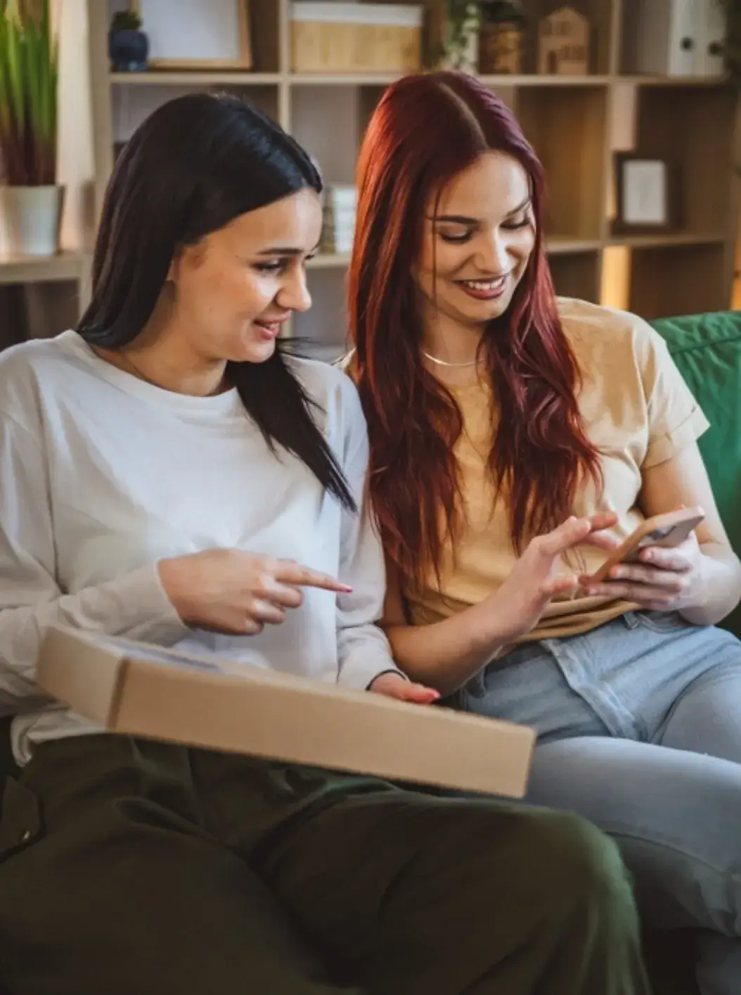 Two smiling friends on a couch as one refers the other to Well Done Cleaning Services on her phone
